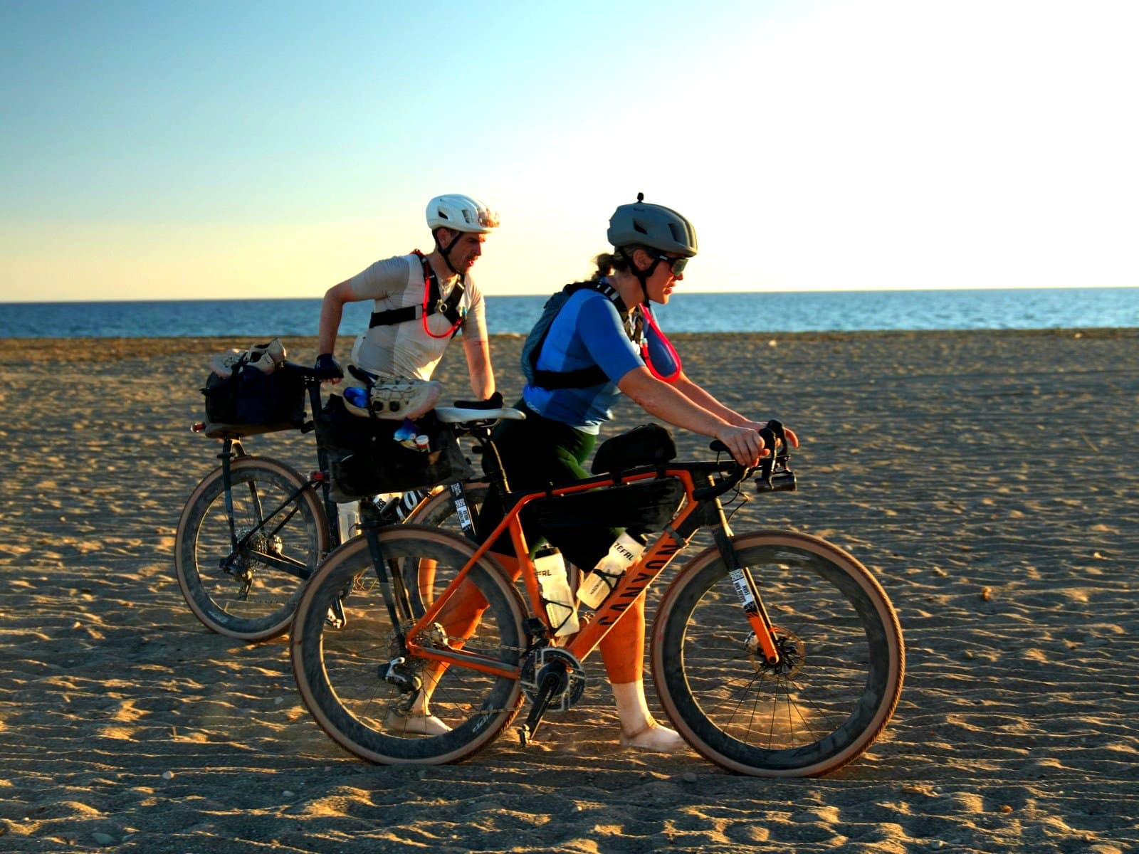 Two riders pushing bikes across a beach at sunset